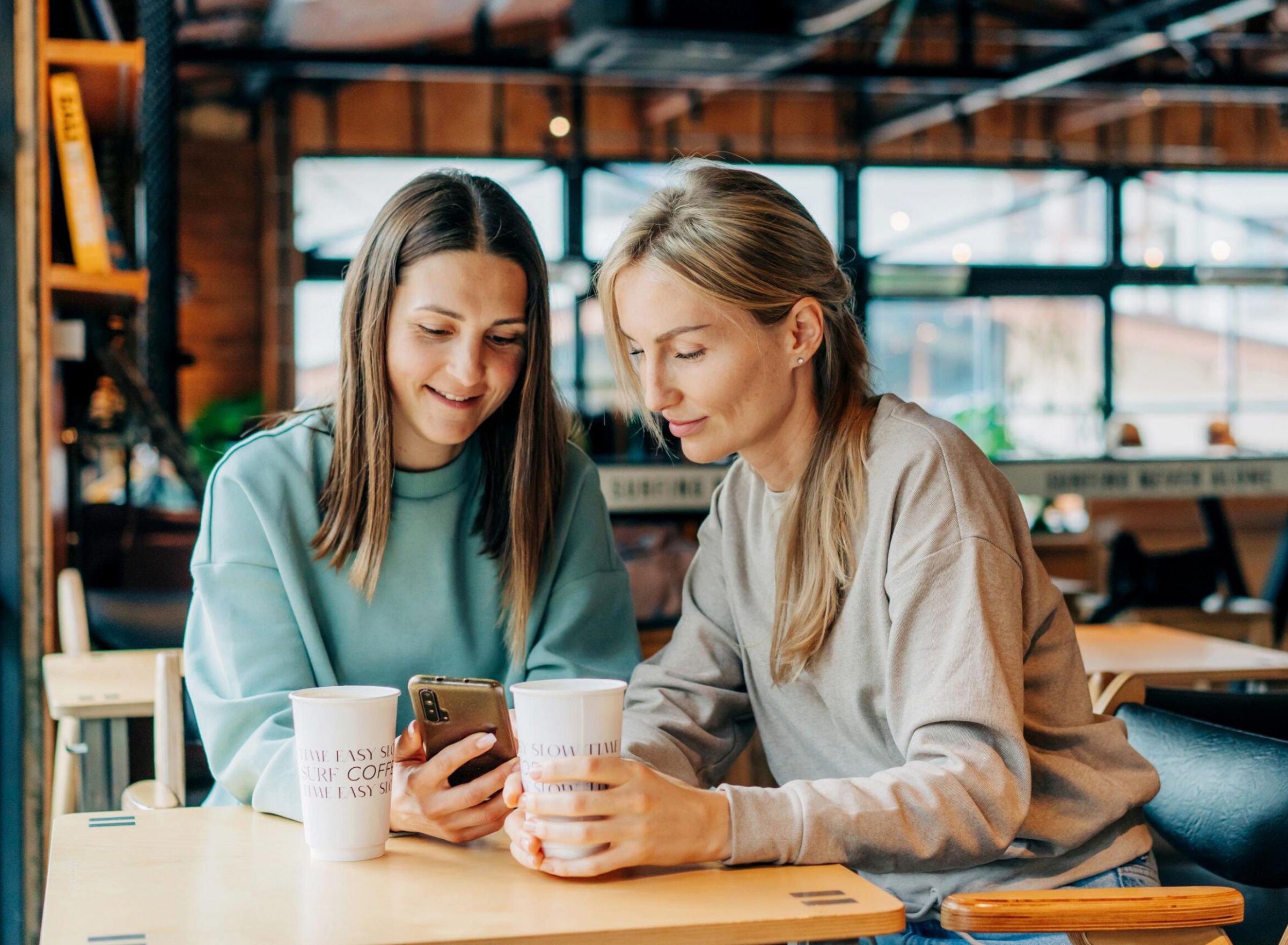 Two women sitting at a cafe table, looking at a phone and holding coffee cups, smiling.
