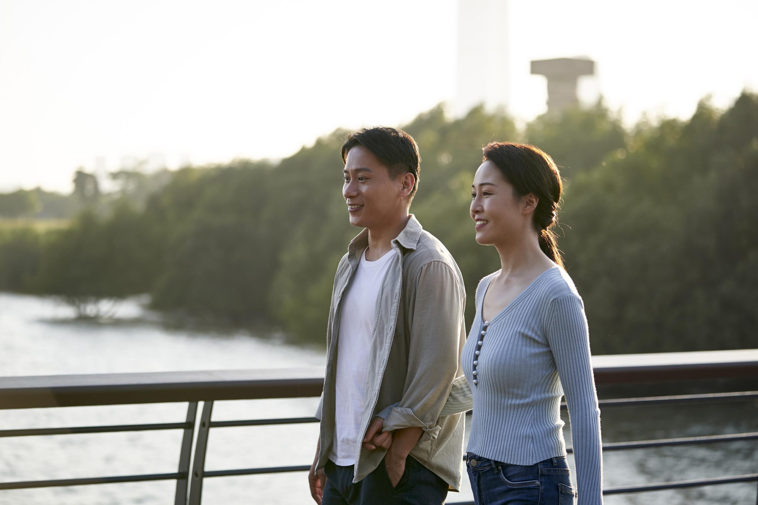 A smiling couple walks together on a riverside bridge with trees in the background.
