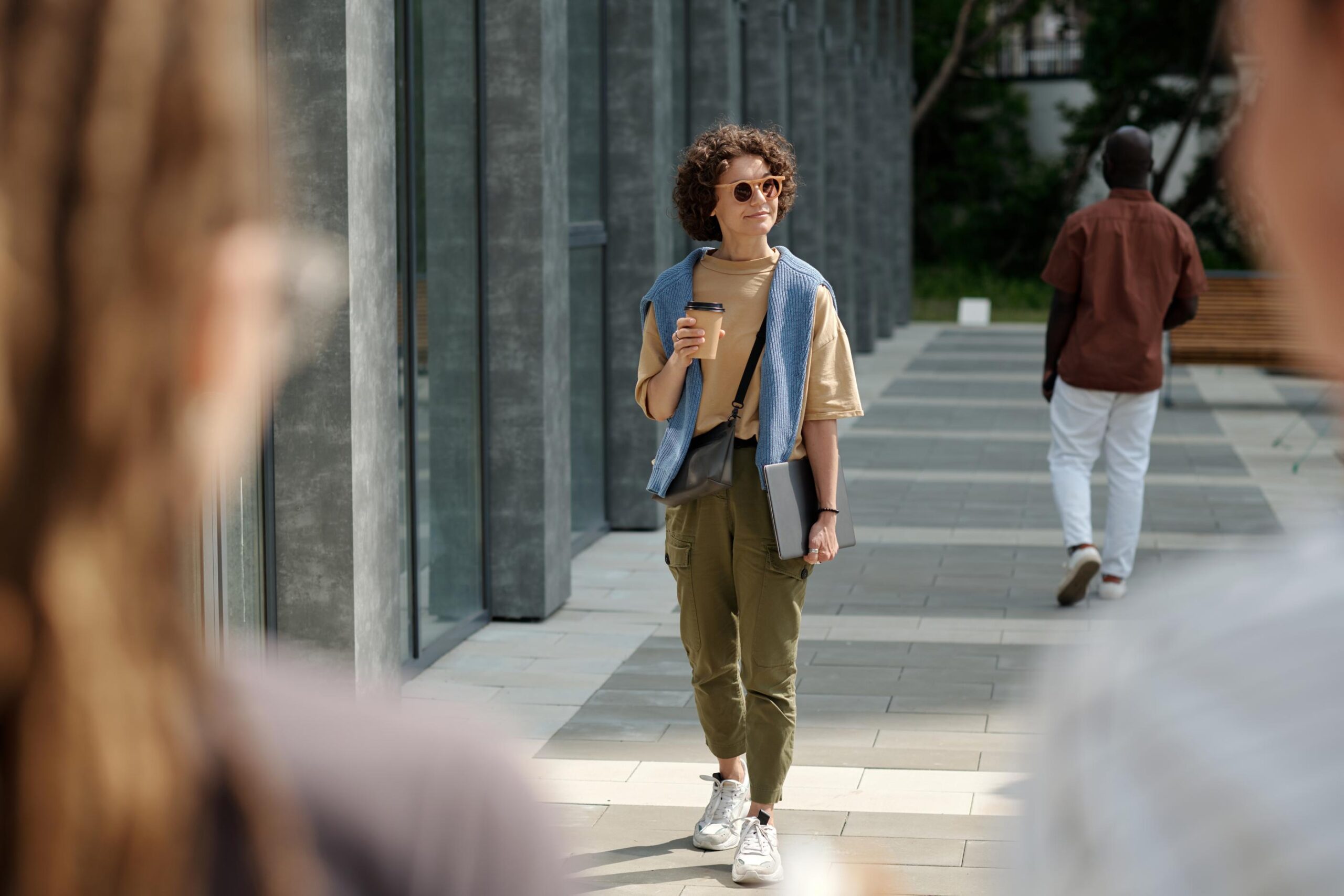 Person with curly hair walking outside, holding a coffee cup and a laptop, wearing sunglasses and casual clothes.